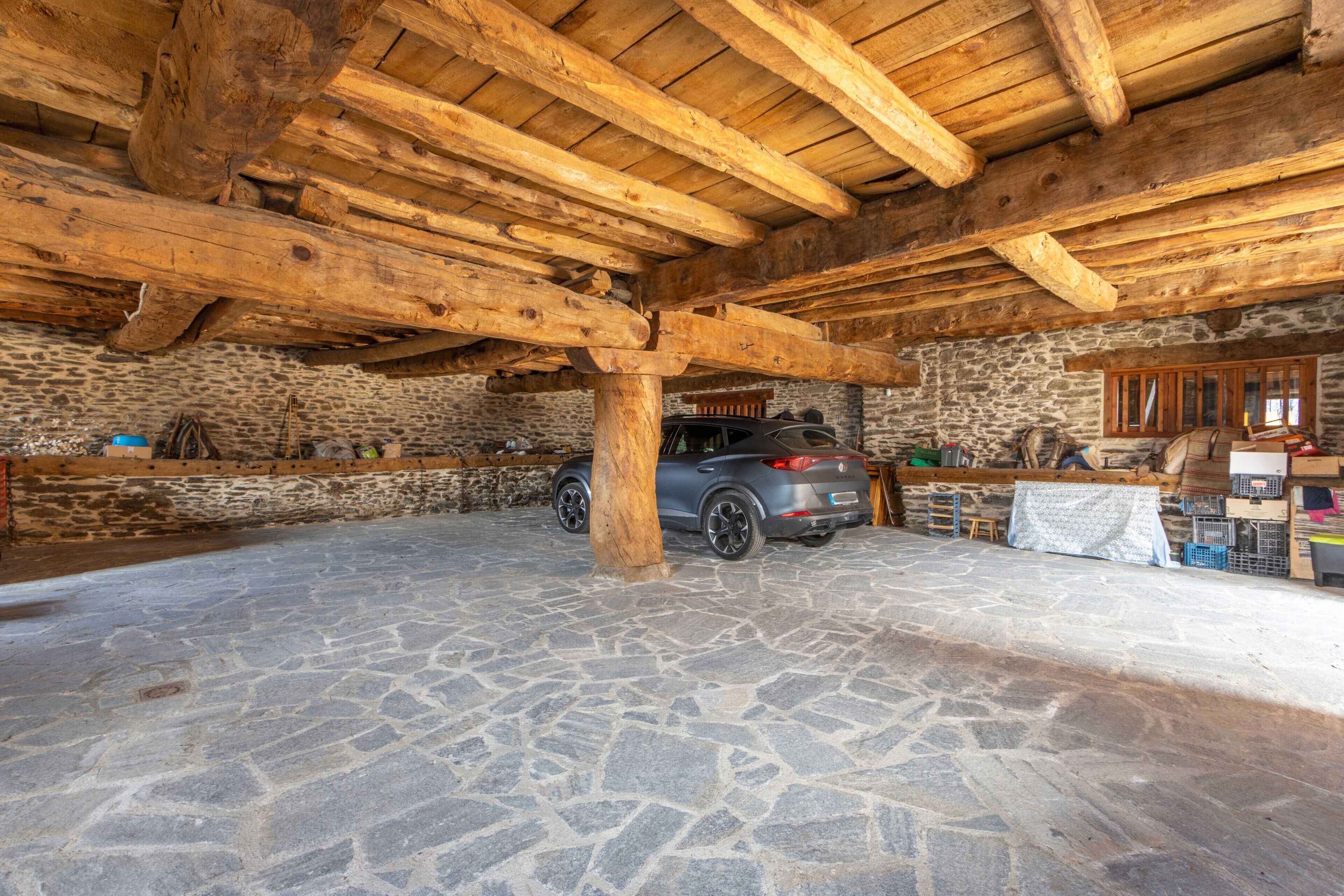 Una habitación con piso de piedra y un coche estacionado bajo una estructura de vigas de madera.