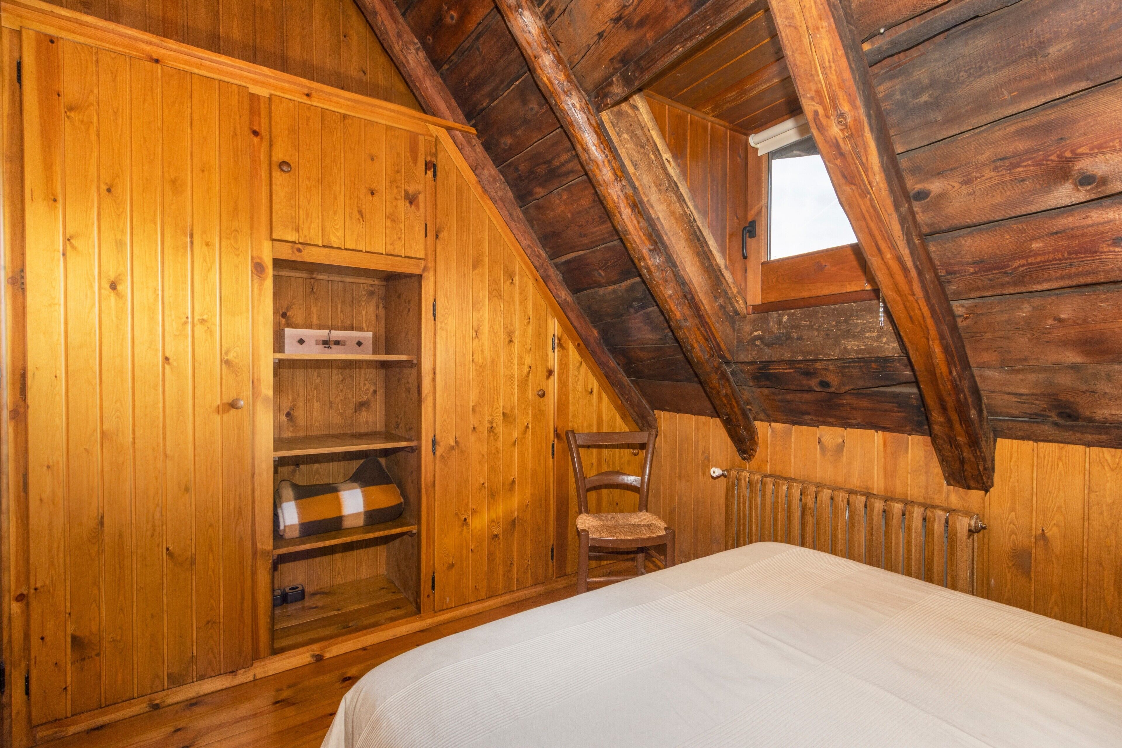 Una habitación de madera con techo inclinado, una cama, una silla y estanterías empotradas.