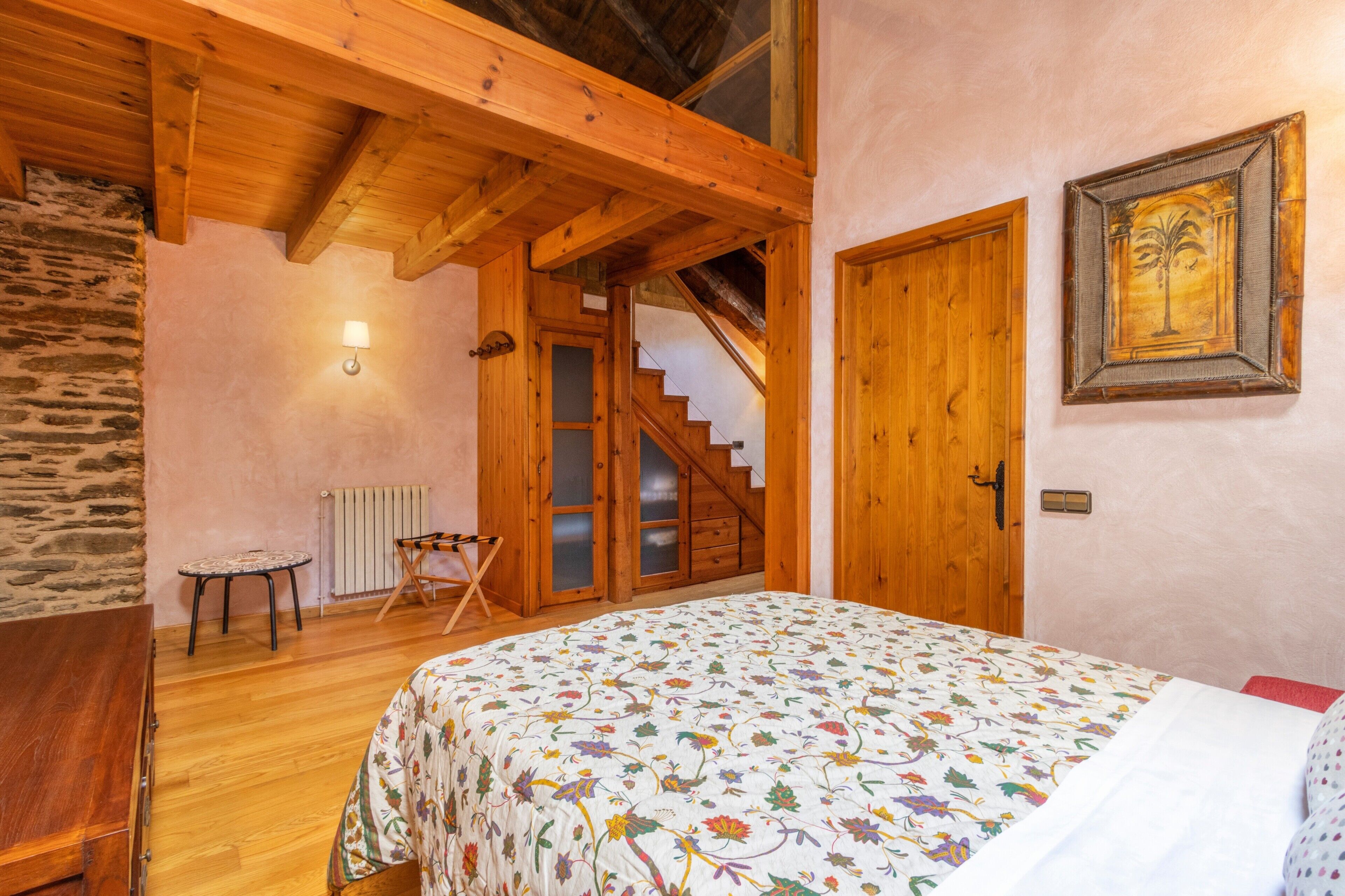 A bedroom with a wooden ceiling, a bed with a floral bedspread, a wooden door, a staircase, and a stone wall.