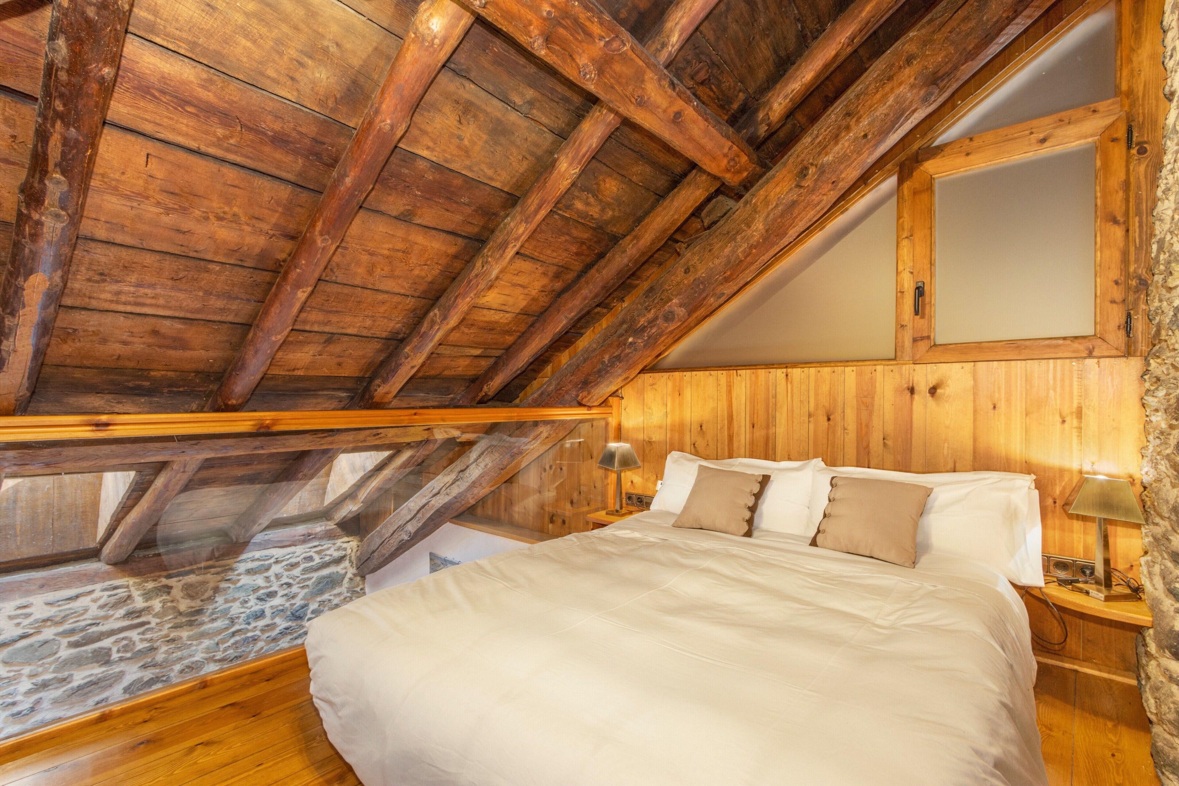 A bedroom with a sloped ceiling, wooden beams, a stone floor, a bed with white linens, a nightstand with a lamp, and a wooden-framed mirror.