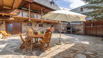 A stone patio with a wooden table and chairs, a large umbrella, and a stone building with wooden balconies.