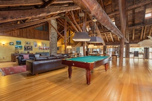 Um quarto amplo com mesa de bilhar, sofá de couro, vigas de madeira e um pilar de pedra.