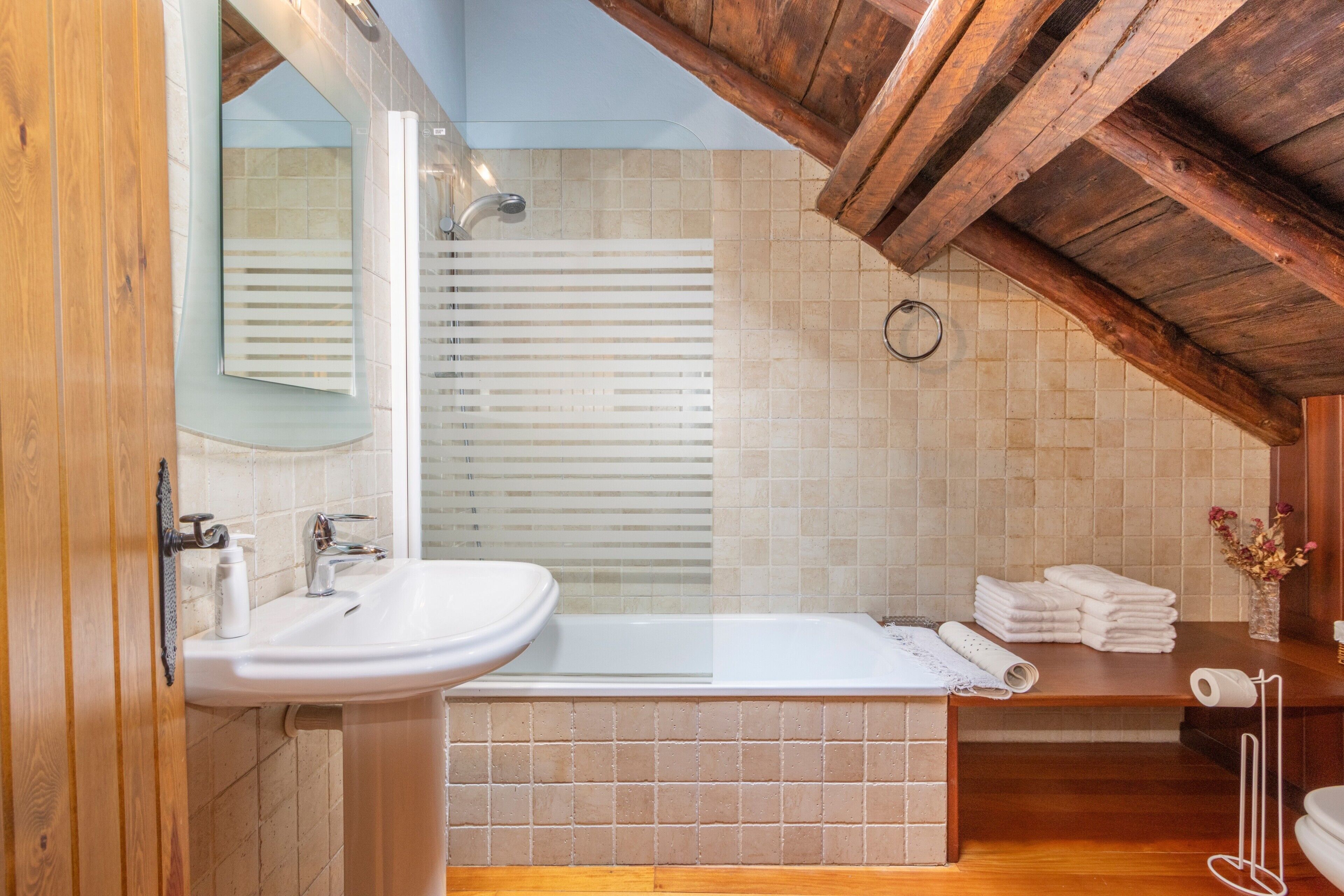 A bathroom with a sink, mirror, bathtub, wooden ceiling, and tiled walls.