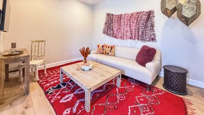 Un salon avec une table basse en bois, un canapé blanc, un tapis à motifs rouges, un ventilateur de plafond et des décorations murales hexagonales.