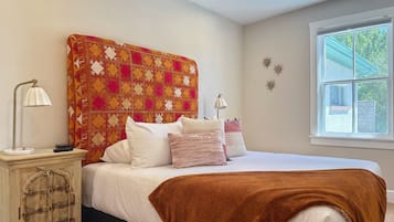 Un dormitorio con cama, almohadas, una manta, una mesita de noche, una lámpara y una ventana.