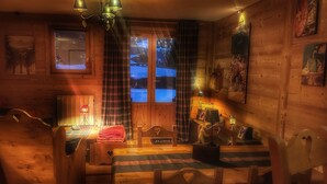 L’intérieur chaleureux d’un chalet, avec un revêtement mural en bois, un lit recouvert d’une couverture à carreaux, une table avec des chaises et une fenêtre agrémentée de rideaux.