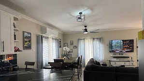 Eine moderne Küche mit weißen Schränken, einer schwarzen Arbeitsplatte und einem Deckenventilator. Es gibt einen Essbereich mit Tisch und Stühlen sowie ein Wohnzimmer mit einer schwarzen Couch und einem Fernseher.