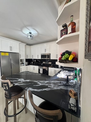A modern kitchen with white cabinets, a stainless steel refrigerator, a microwave, and a gas stove. There is a black countertop with a marble-like pattern, two wooden stools, and a fruit basket on a shelf. - Jahiem's Paradise (Ocho Rios)
