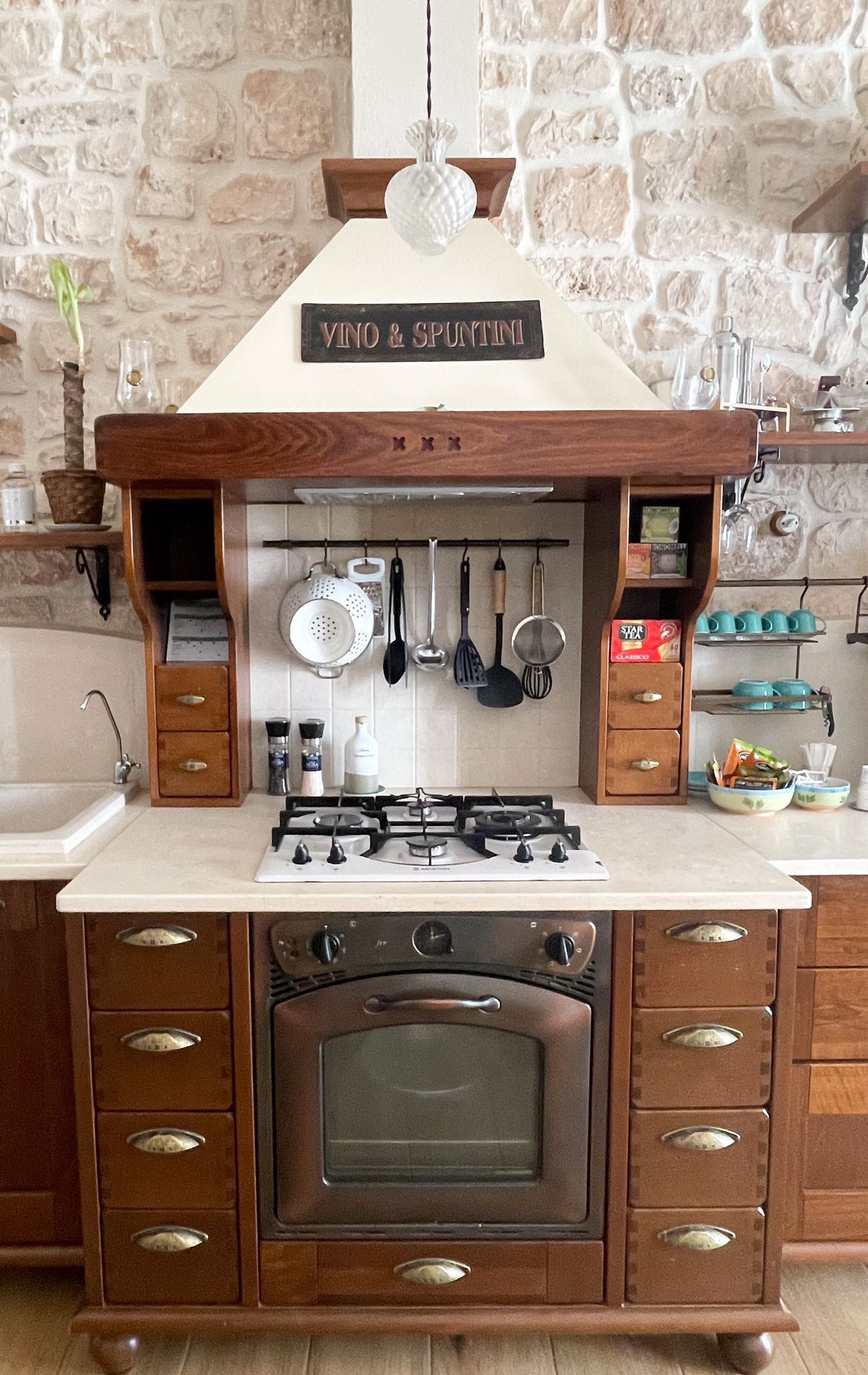 Una cocina con pared de piedra, un mueble de madera con cajones, una estufa a gas y utensilios colgantes.