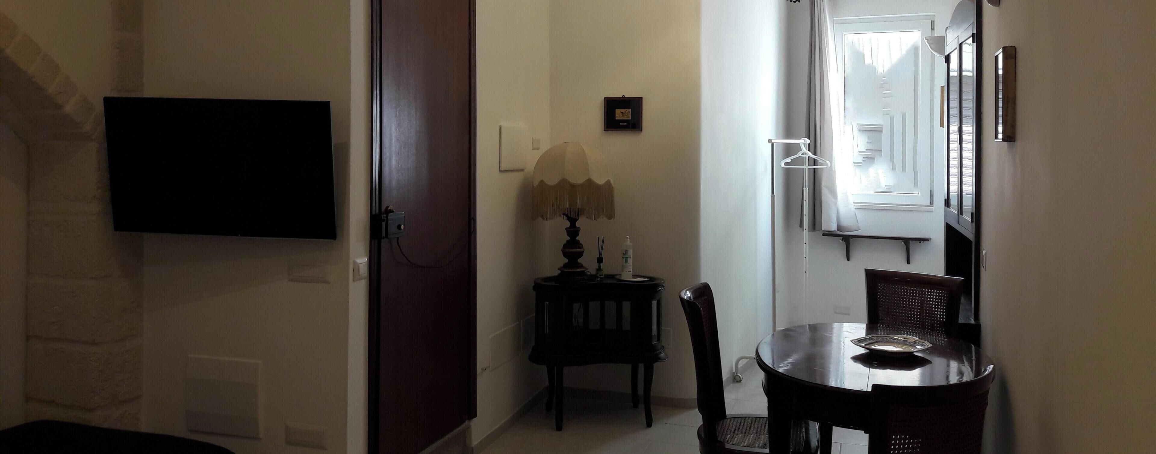 Un pasillo con una televisión montada en la pared, una puerta, una mesita con una lámpara y un comedor con sillas y una mesa.