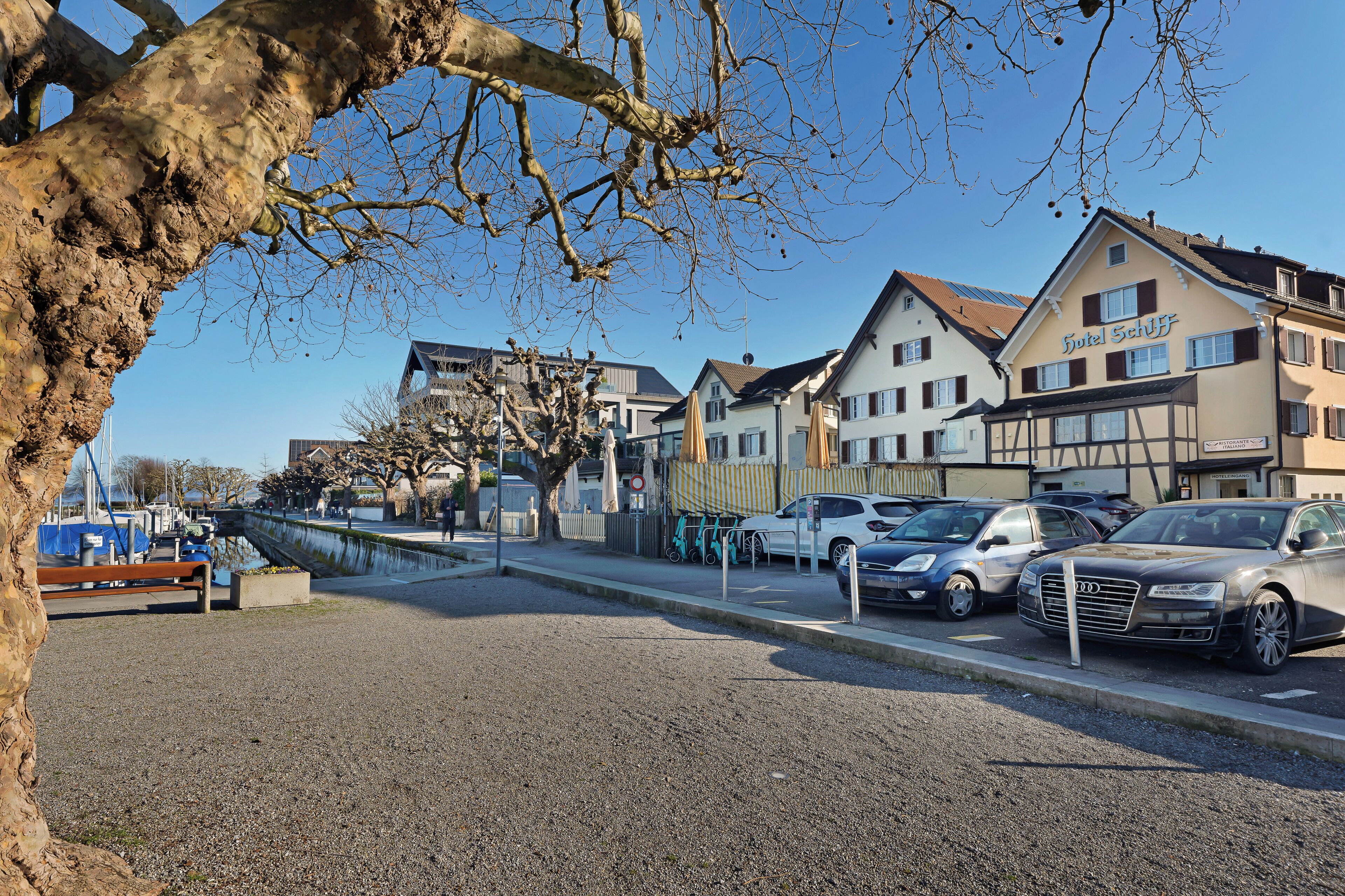 A hotel with a traditional European facade, a parking area with cars, and a tree in the foreground.