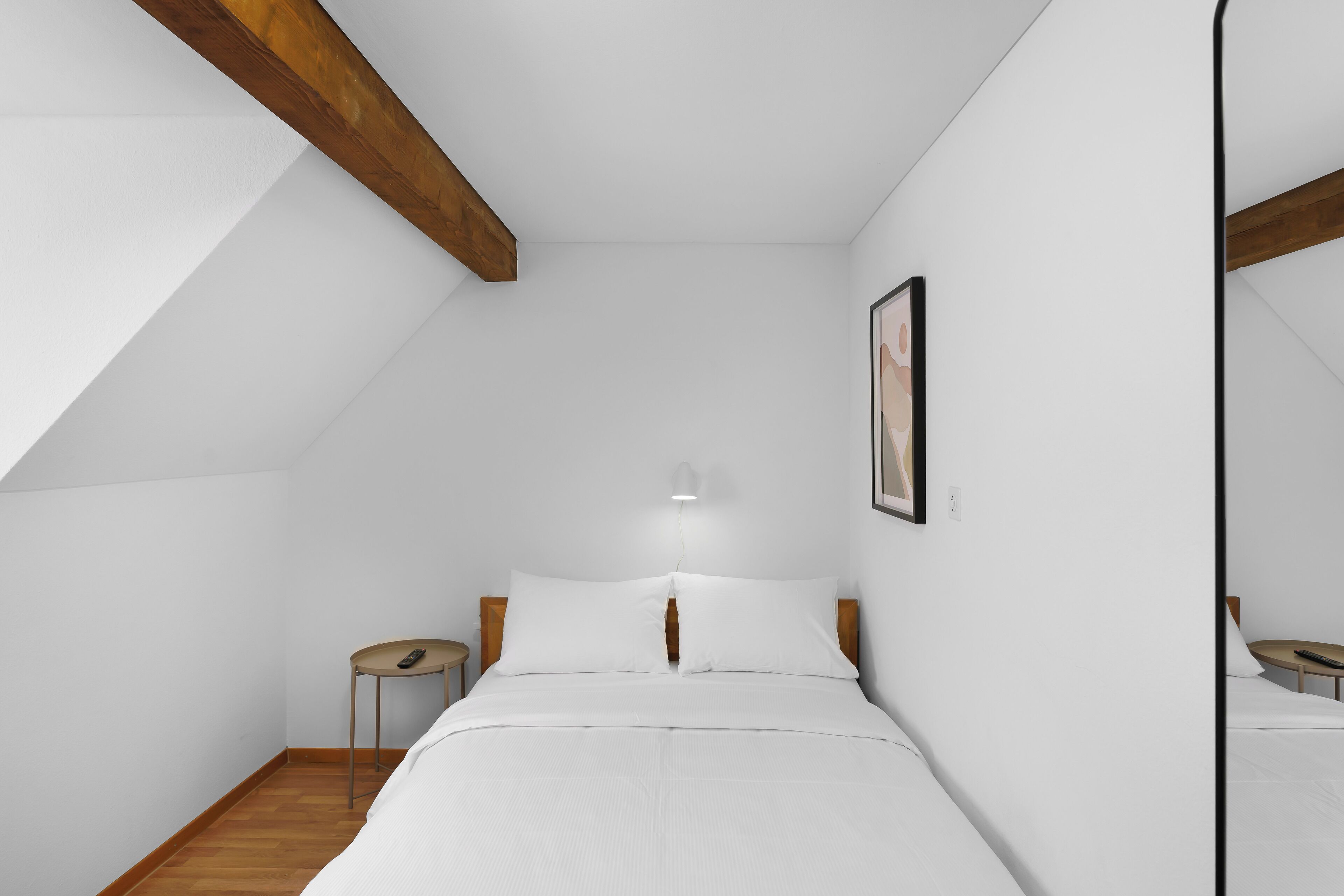A bedroom with a sloped ceiling, a bed with white bedding, a bedside table with a lamp, and a framed picture on the wall.