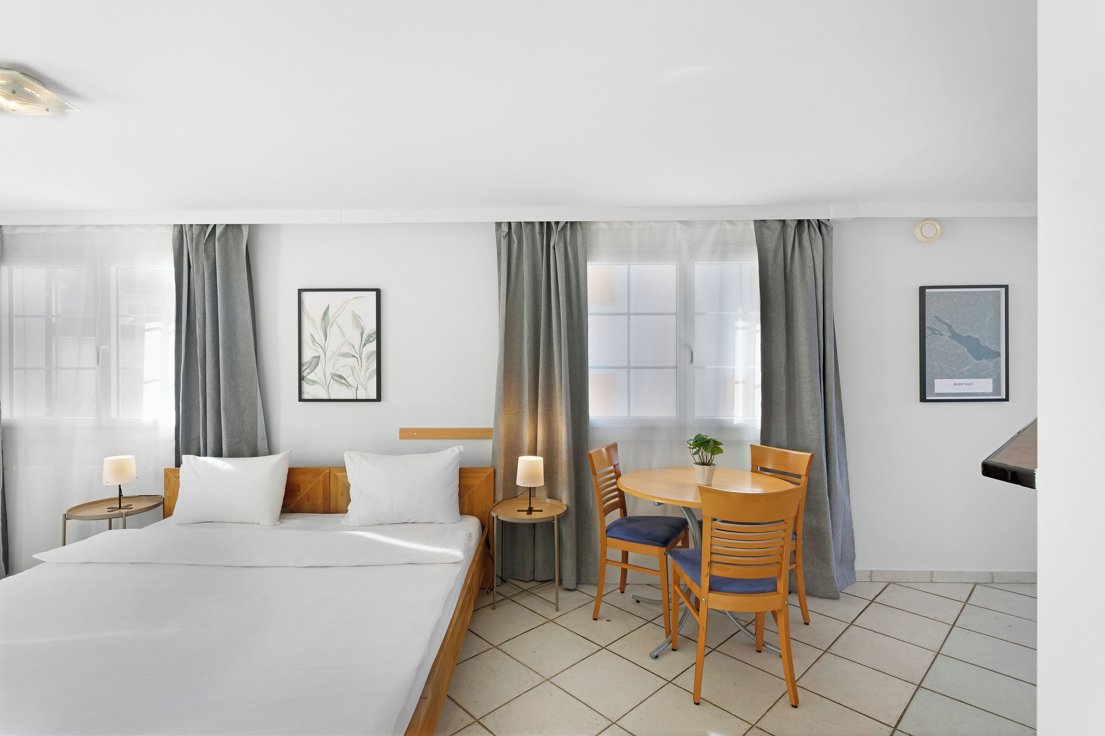 A hotel room with a large bed, a small dining table with chairs, a lamp, and two framed pictures on the walls.