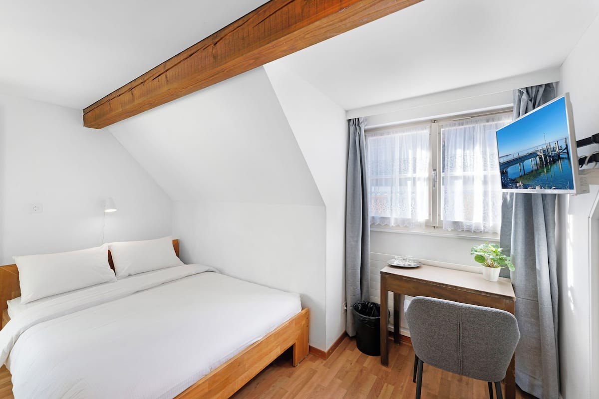 Ein Schlafzimmer mit Holzbalkendecke, einem Bett, einem Schreibtisch mit Computer, einem Stuhl und einem Fenster mit Vorhängen.