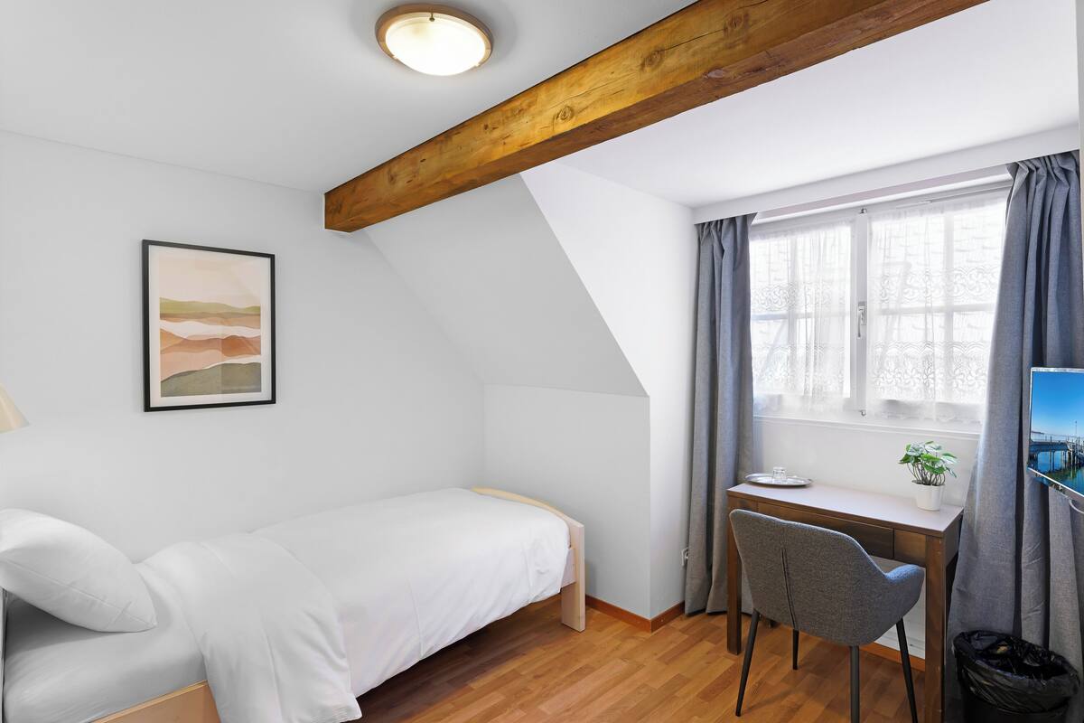 Ein Schlafzimmer mit Bett, Schreibtisch, Stuhl, einem Fenster mit Vorhängen, einem Bild an der Wand und einer Deckenleuchte.