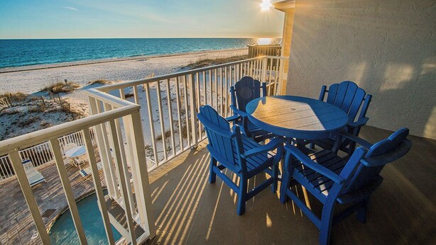 A balcony with a view of the beach and ocean, a round table with blue chairs, and a pool area. - Shoreline Towers (Gulf Shores)