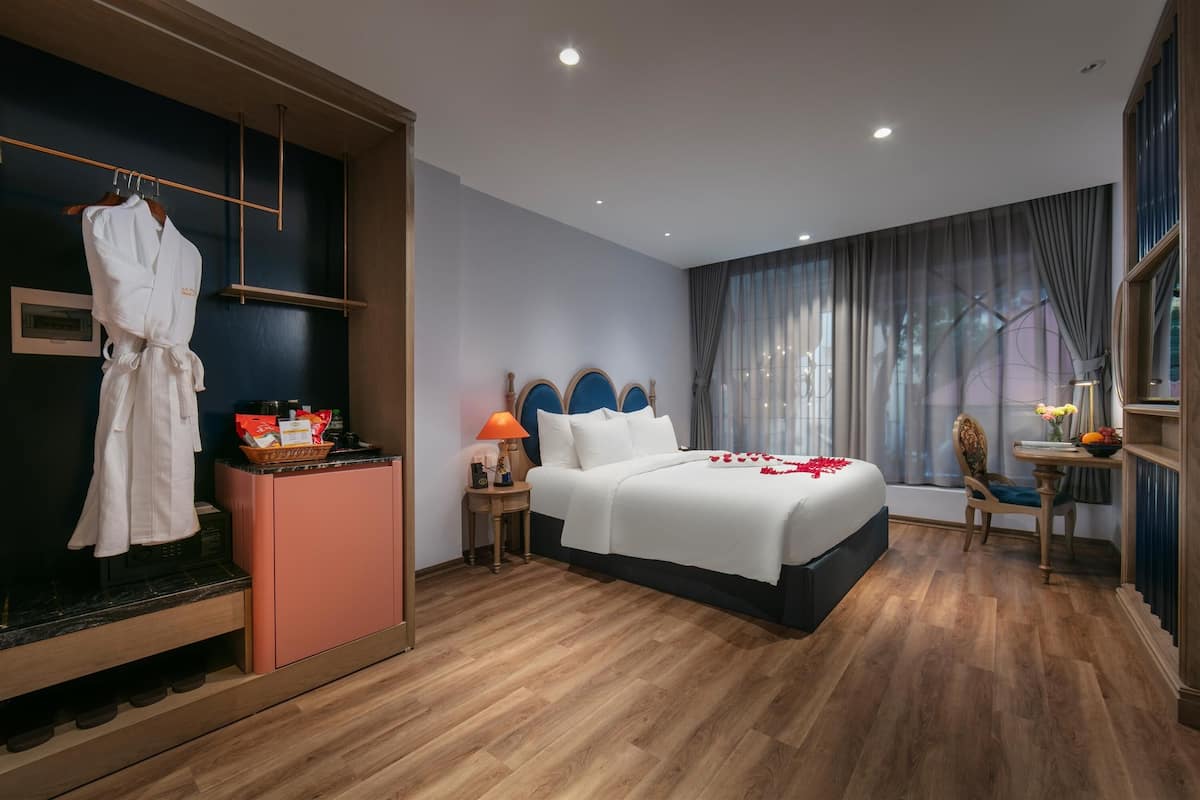 A hotel room with a bed, a side table with a lamp, a chair, a desk with a vase, a robe hanging, and a shoe rack.