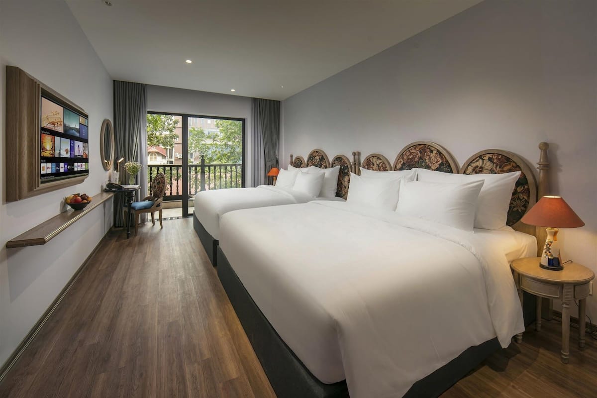 A hotel room with a large bed, a TV mounted on the wall, a small table with a lamp, and a balcony with curtains.