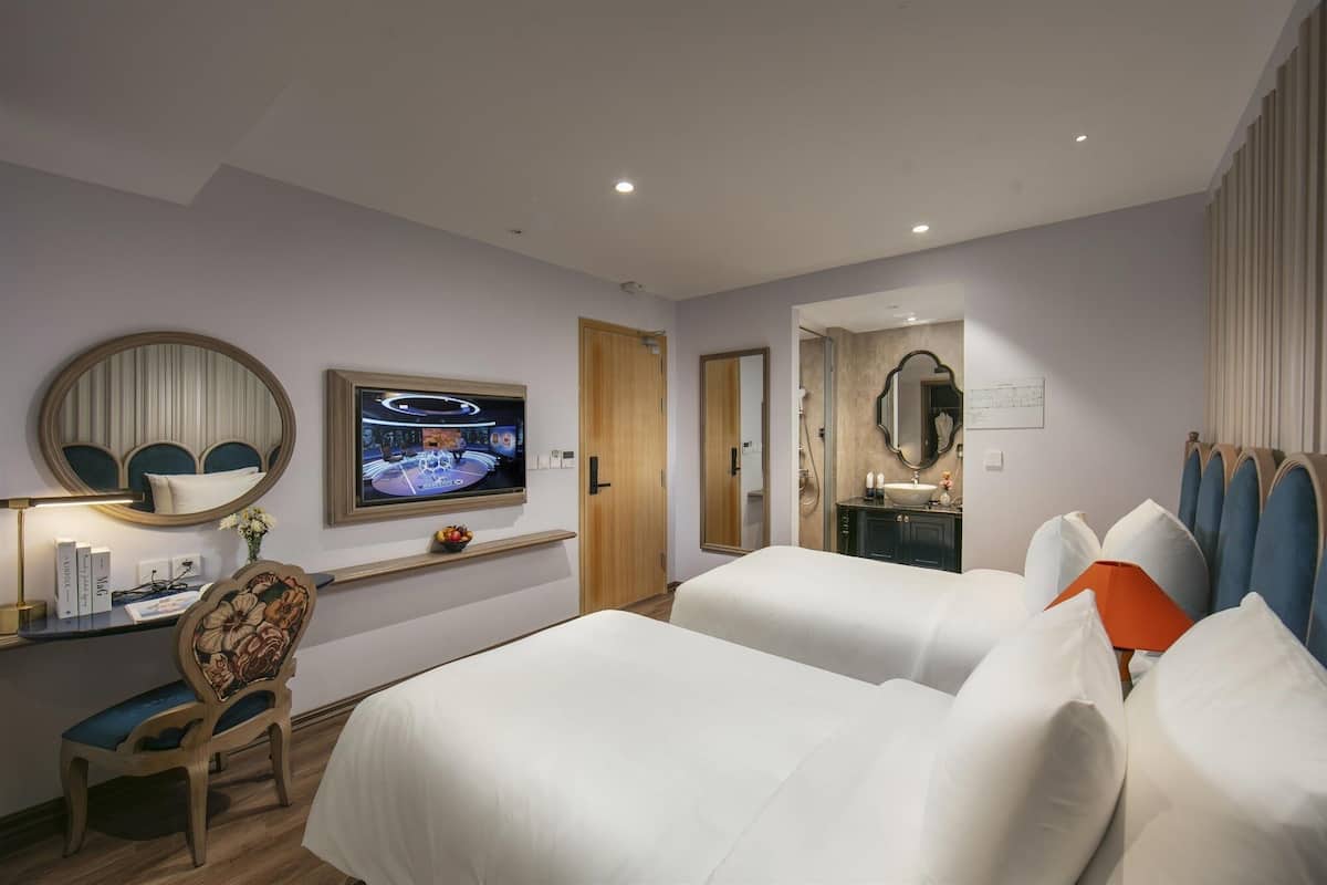 A hotel room with a bed, a desk with a chair, a television, a mirror, and a bathroom visible through an open door.