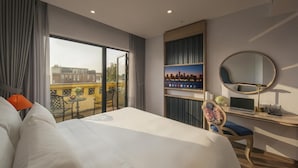 Une chambre d’hôtel avec un lit, un bureau avec un ordinateur portable, une chaise, un grand miroir rond et une vue sur les immeubles à travers de grandes fenêtres.