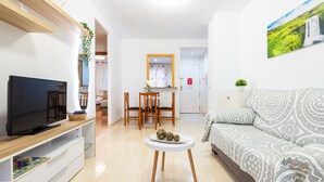 A modern living room with a flat-screen TV, a sofa, a dining table with chairs, and a kitchen area in the background. - Seafront Apartment 'Vistamar' with Sea Views, Wi-Fi & Air Conditioning (Oropesa)