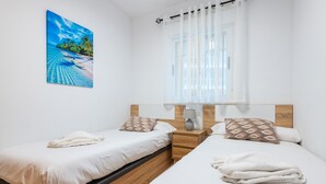 A hotel room with two beds, a window with curtains, a painting of a beach, and a bedside table with a lamp. - Seafront Apartment 'Vistamar' with Sea Views, Wi-Fi & Air Conditioning (Oropesa)