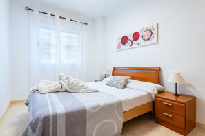 Una cama bien hecha con cabecera, una mesita de noche con lámpara, y una obra de arte colgada en la pared.