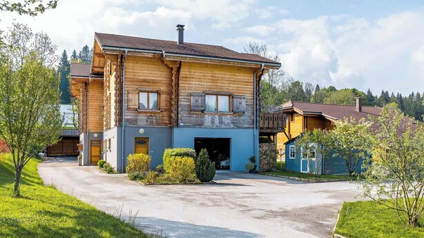 Une maison en bois à deux étages, dotée d’un garage, entourée de verdure et baignée par un ciel dégagé.