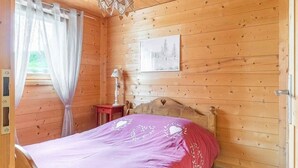 Une chambre dans une cabane en bois, avec un lit, une table de chevet, une lampe, une fenêtre avec des rideaux, un luminaire au plafond et un tableau encadré accroché au mur.