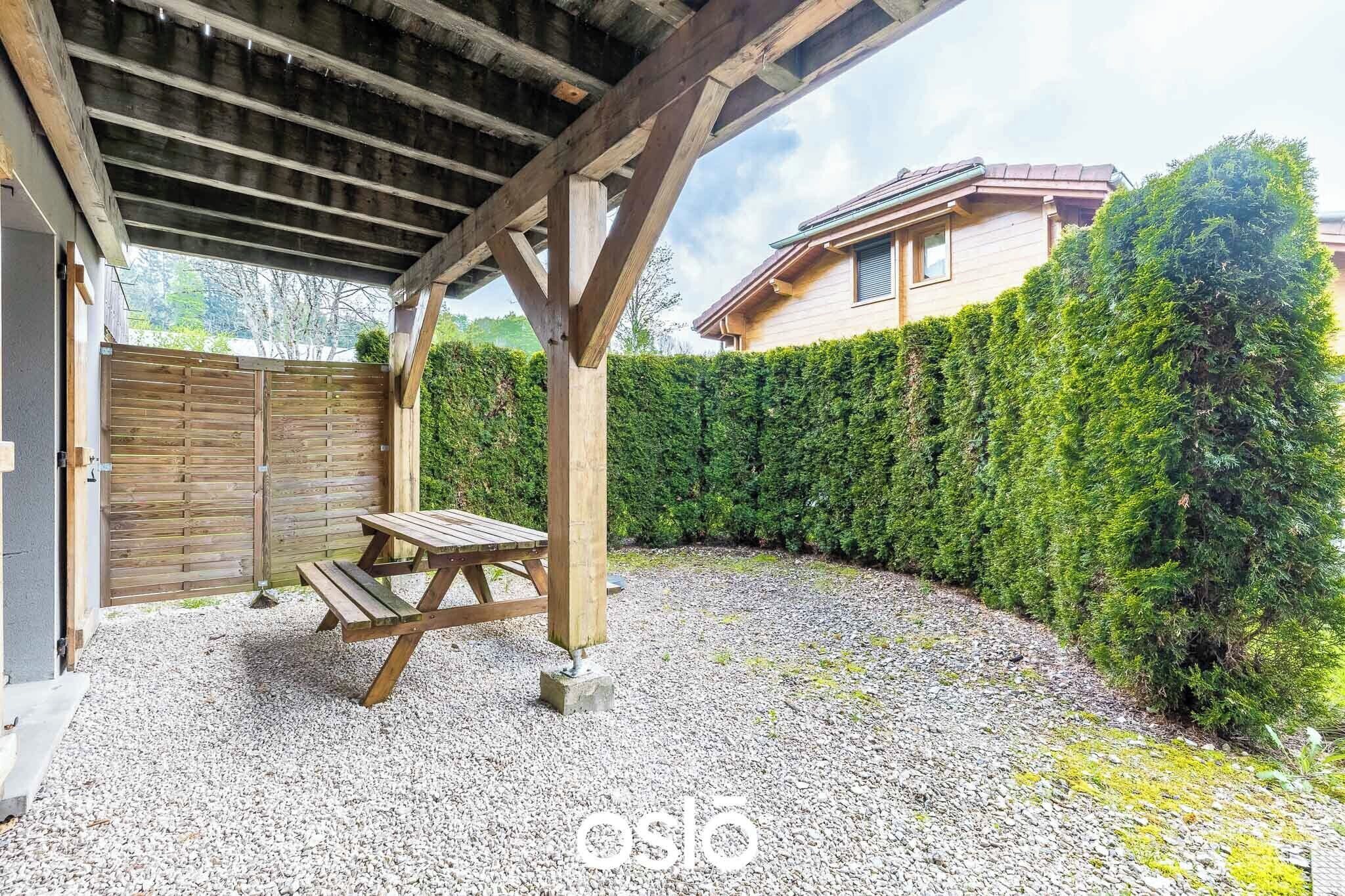 A covered outdoor area with a wooden table and benches, a gravel ground, a wooden fence, and a hedge.