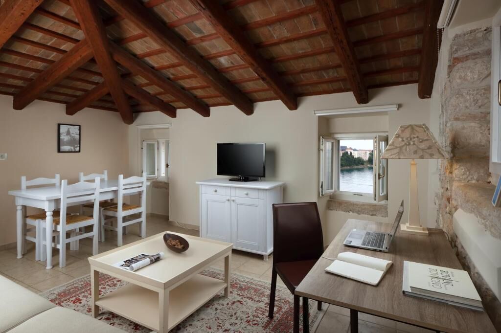 Een knus vertrek met een houten plafond, een televisie op een witte kast, een eethoek met witte stoelen, een tafel met een schaal en tijdschrift, en uitzicht op een waterpartij door een raam.