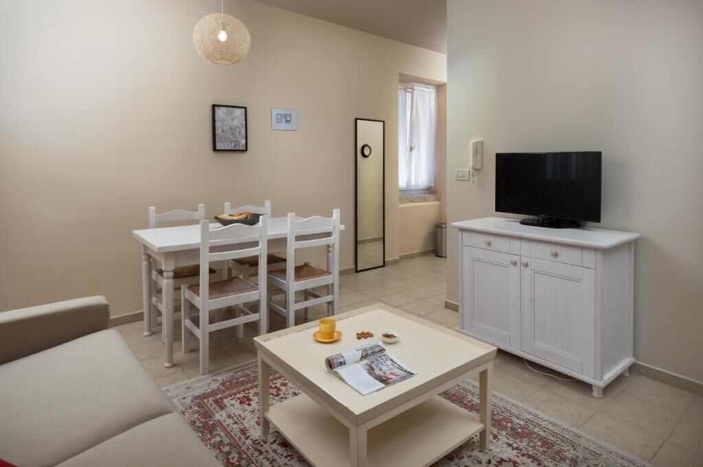 A small, well-lit room with a white dining table and chairs, a television on a cabinet, and a large mirror on the wall.