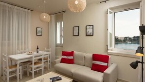 Ein modernes Wohnzimmer mit einem weißen Sofa, roten Kissen, einem Esstisch mit Stühlen und einem großen Fenster mit Blick auf ein Gewässer.