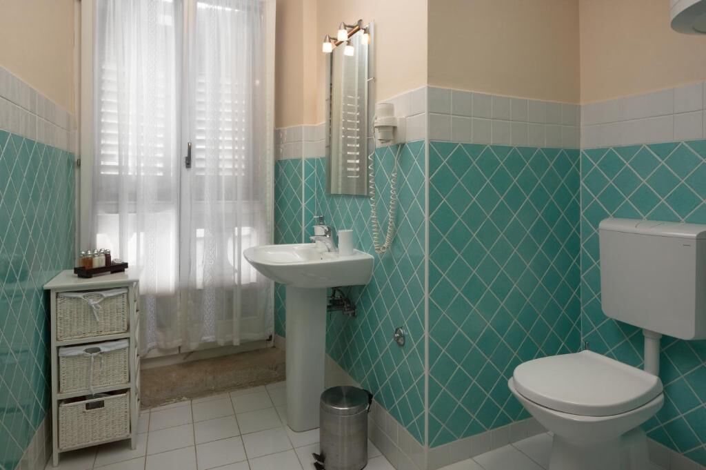 Ein Badezimmer mit Waschbecken, Toilette und Spiegel. Ein Fenster mit Vorhang und einem Mülleimer sind ebenfalls vorhanden.