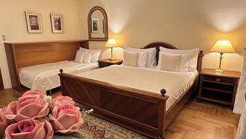 Camera d'albergo con due letti singoli e un letto matrimoniale, testiere in legno, comodini con lampade, uno specchio sopra il letto matrimoniale e un vaso di fiori sul pavimento.