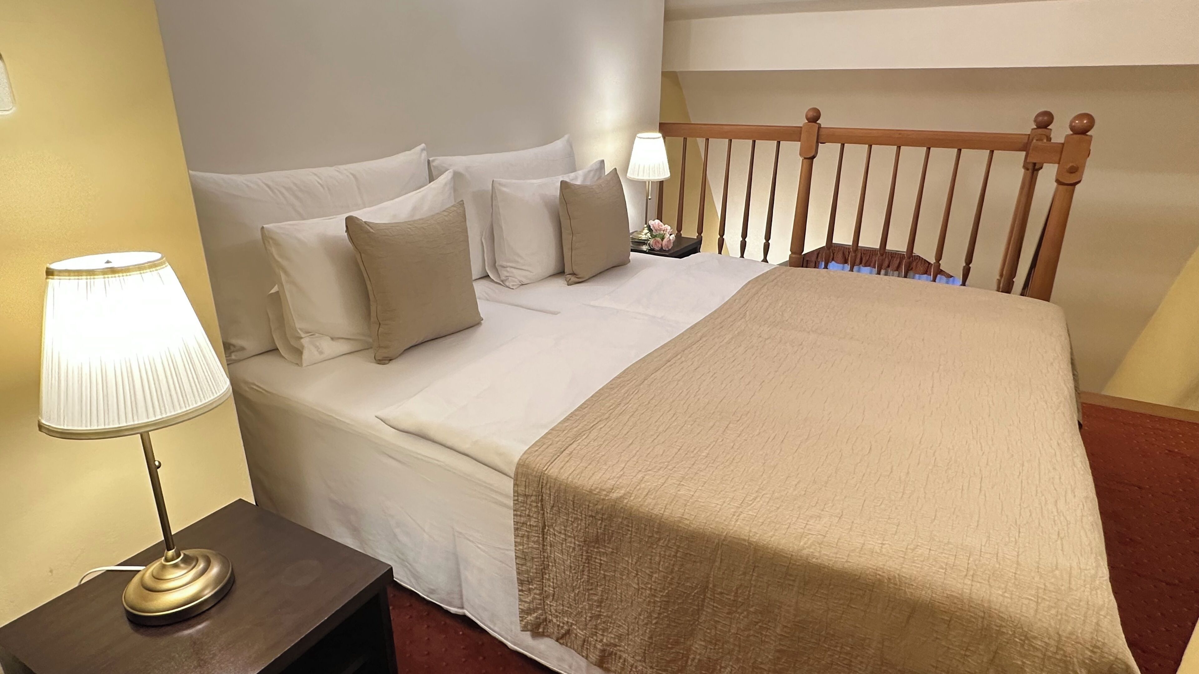 A neatly made bed with white linens and a beige throw blanket, a bedside lamp, and a wooden headboard.