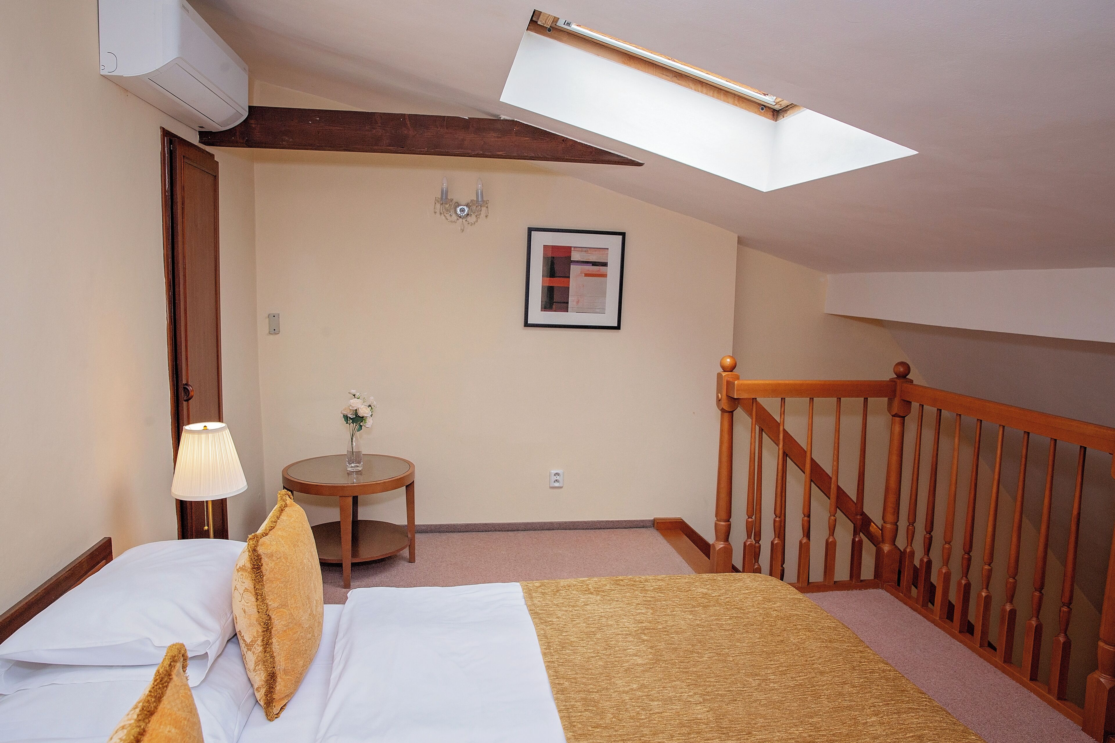 Habitación de hotel con cama, lámpara, mesita con un florero, un cuadro enmarcado en la pared y una escalera de madera.