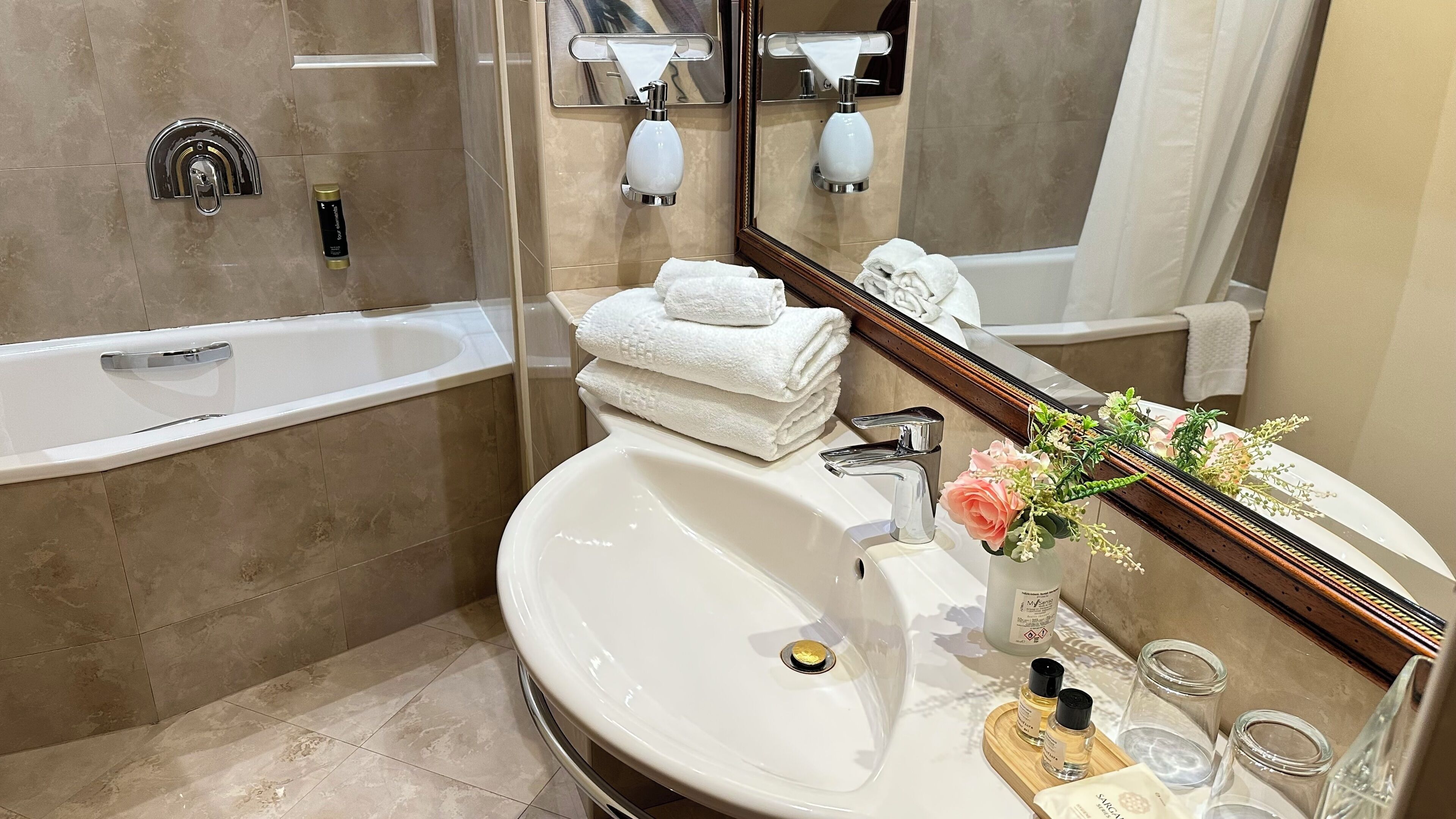 Ein Badezimmer mit einer Badewanne, einem Waschbecken und einem Spiegel. Handtücher liegen auf dem Waschbecken gestapelt, und auf der Ablage steht eine Vase mit Blumen.