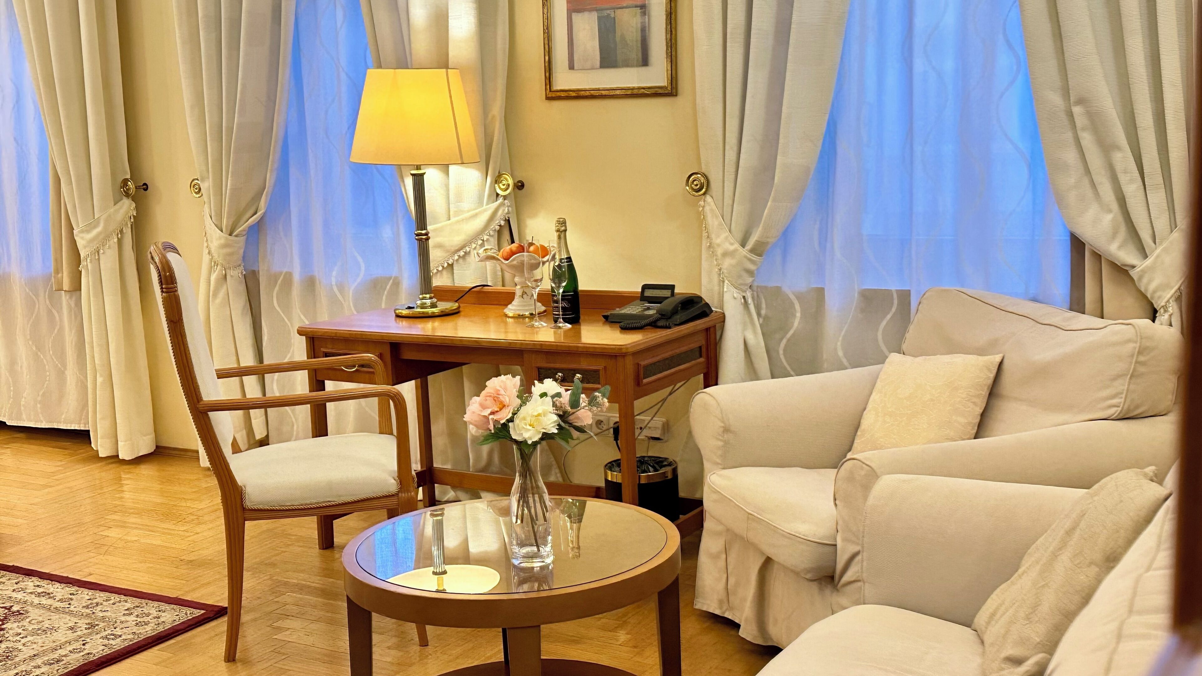 Una habitación de hotel con un escritorio de madera, una silla, un sofá, una mesita redonda con un florero y una lámpara.