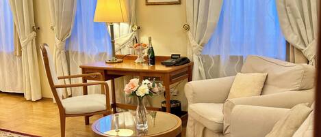 Una habitación de hotel con un escritorio de madera, una silla, un sofá, una mesita redonda con un florero y una lámpara.