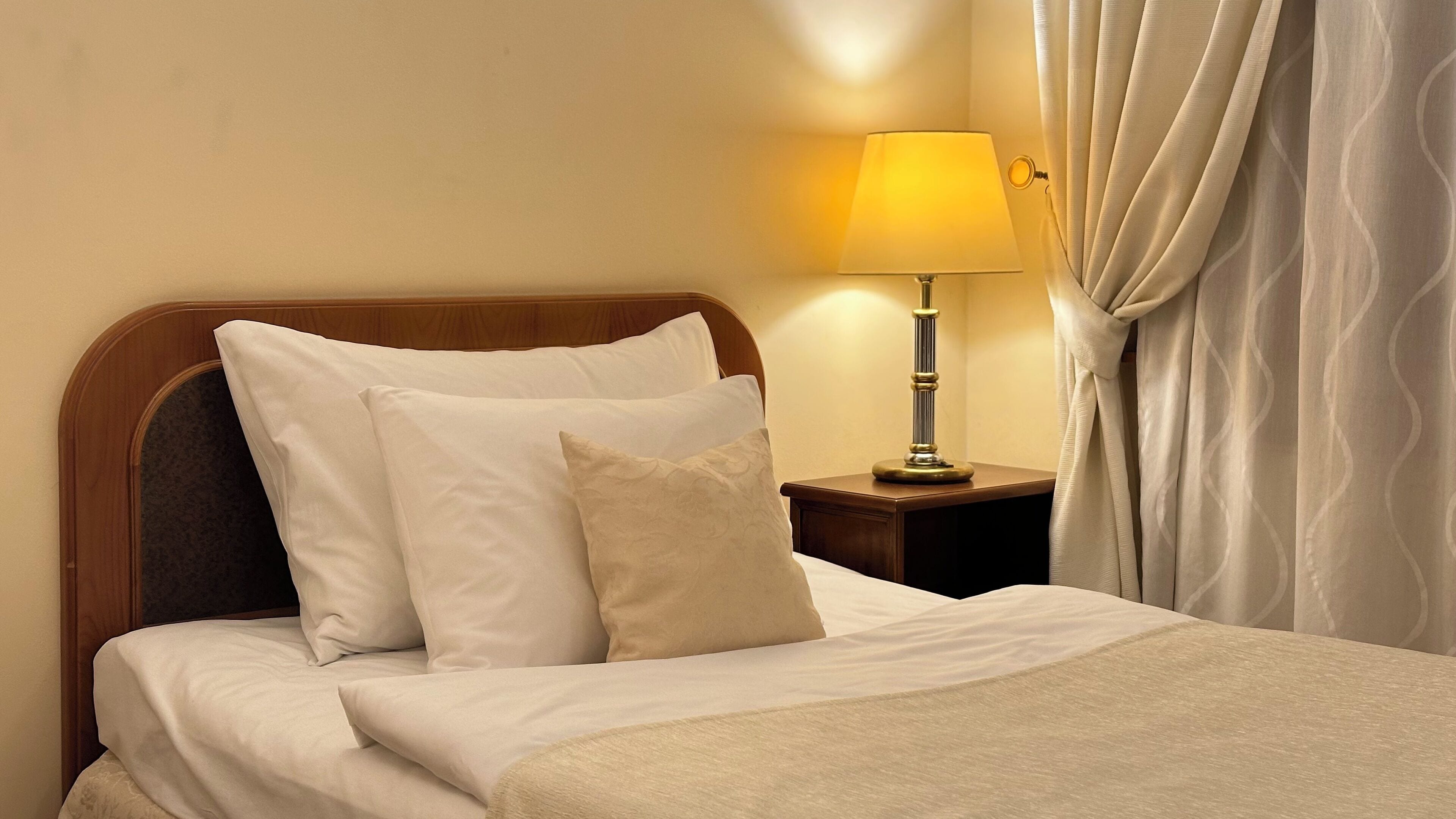 A neatly made bed with white linens and pillows, a bedside table with a lamp, and a curtain partially drawn to the side.