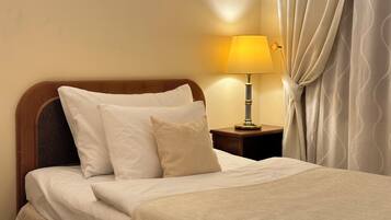A neatly made bed with white linens and pillows, a bedside table with a lamp, and a curtain partially drawn to the side.