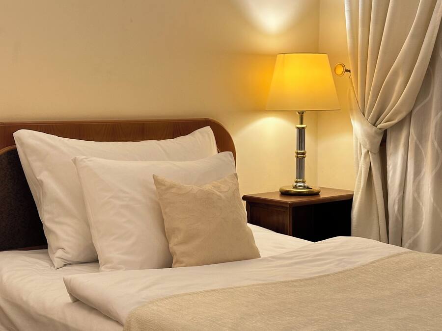 A neatly made bed with white linens and pillows, a bedside table with a lamp, and a curtain partially drawn to the side.