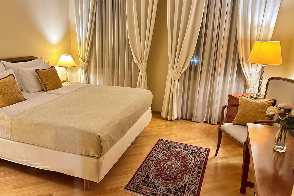 A neatly made bed with a beige bedspread and pillows, a wooden nightstand with a lamp, a wooden chair with a cushion, and a patterned rug on a wooden floor.