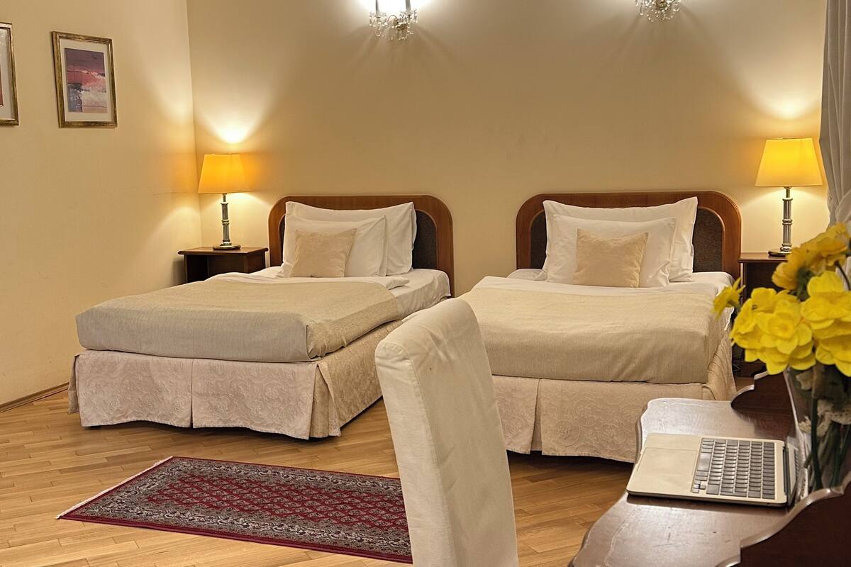 A hotel room with two beds, a desk with a laptop, a red patterned rug, and two lamps.