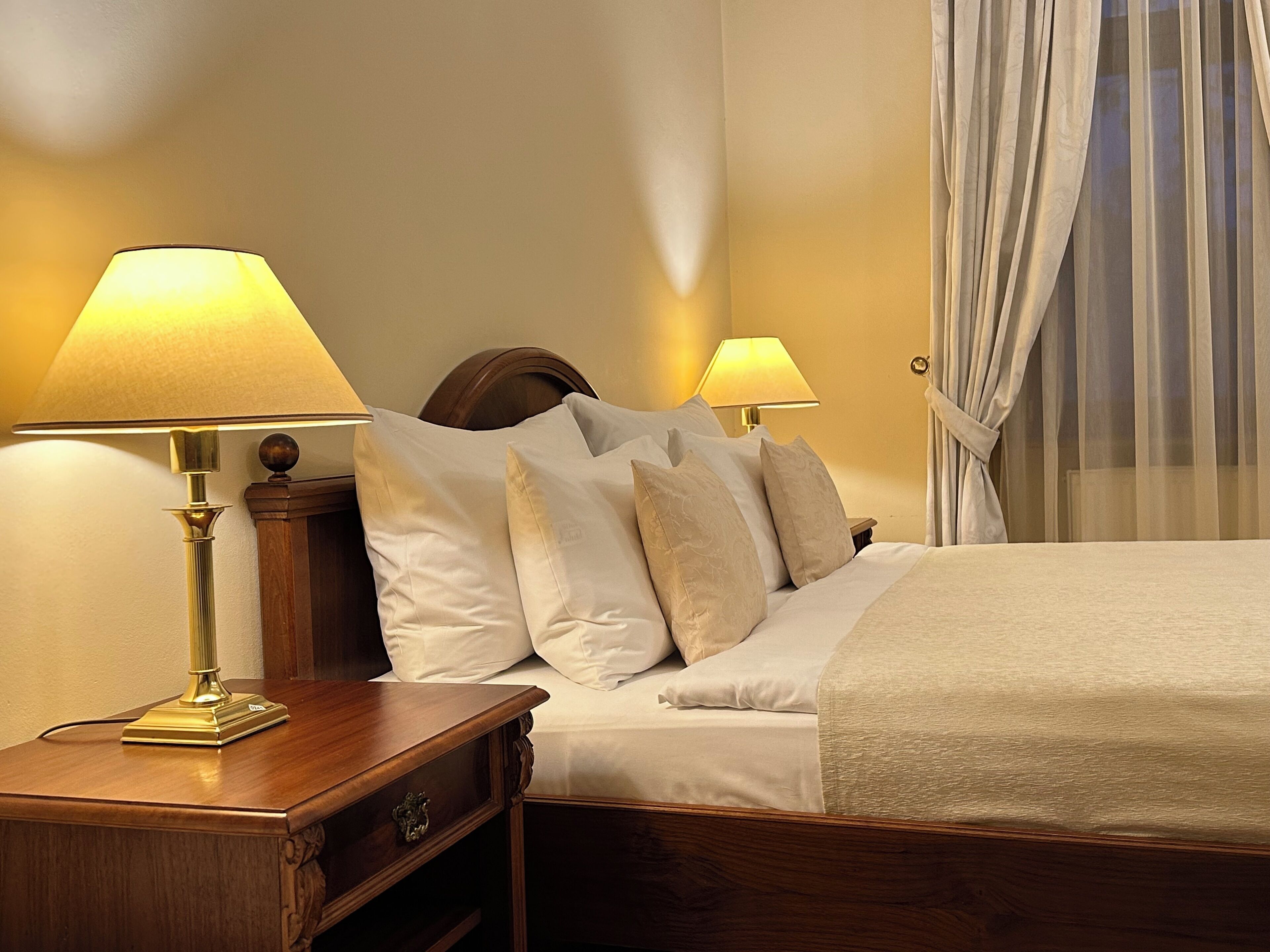 Una cama cuidadosamente tendida con sábanas blancas y varios almohadones, con dos lámparas de noche sobre mesitas de madera.