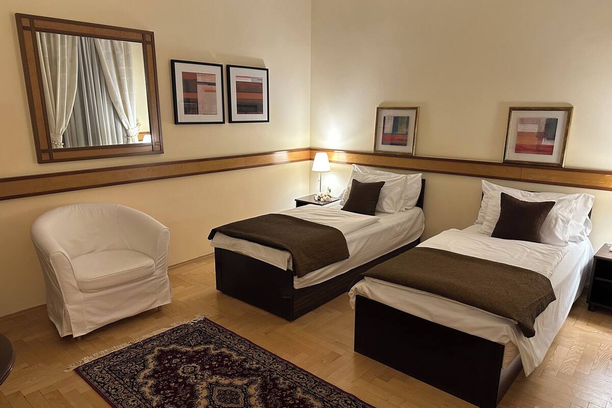 A hotel room with two single beds, a white armchair, a wooden floor, and a patterned rug.