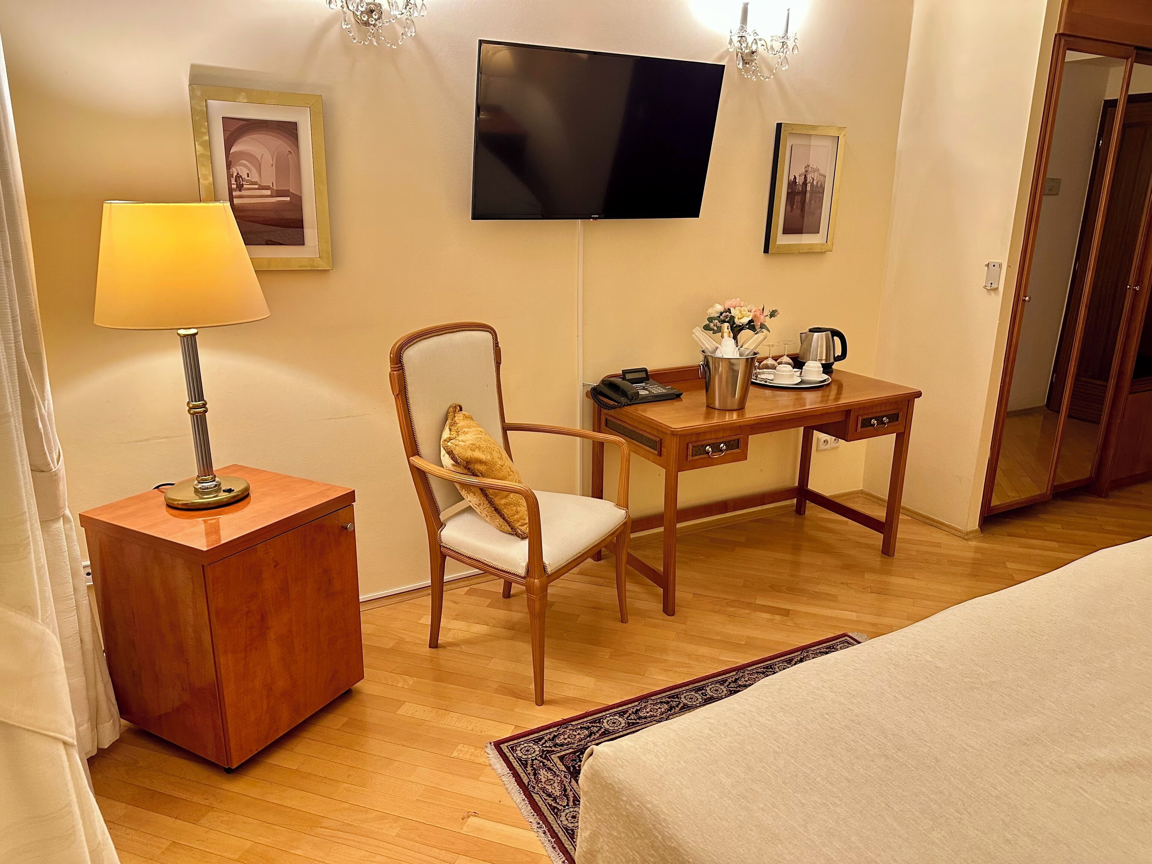 Una habitación de hotel con un escritorio de madera, una silla, un televisor montado en la pared, una lámpara y una cama.