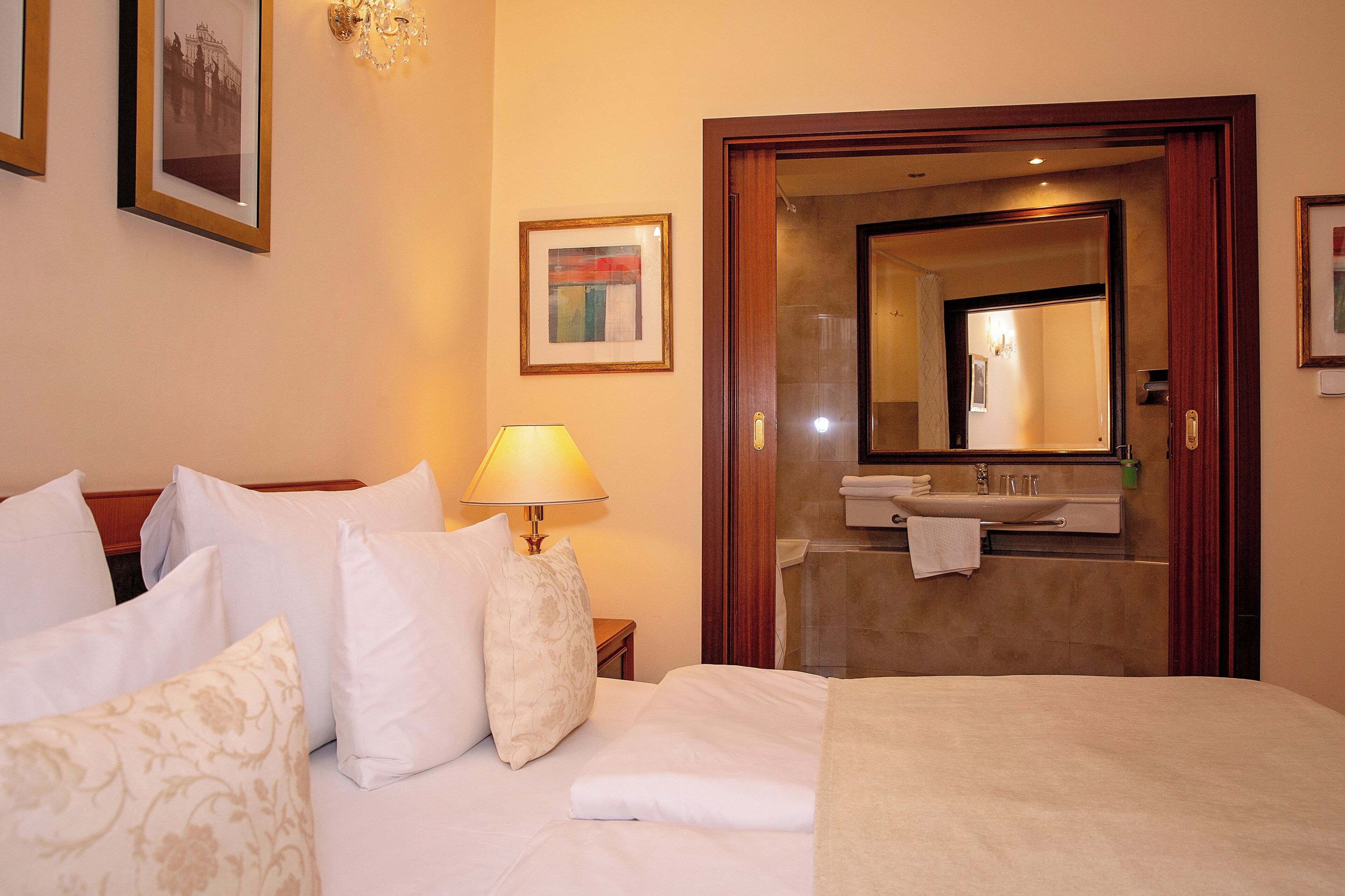 Quarto de hotel com cama, almofadas, abajur, quadro emoldurado e banheiro visível através de uma porta aberta.