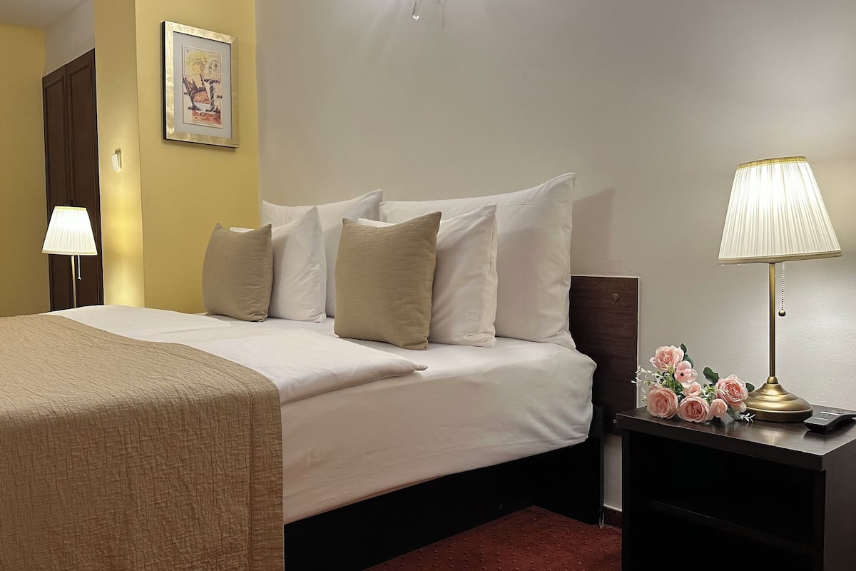 A neatly made bed with a beige blanket, white pillows, and a dark headboard. There is a nightstand with a lamp and a small table with a vase of flowers.